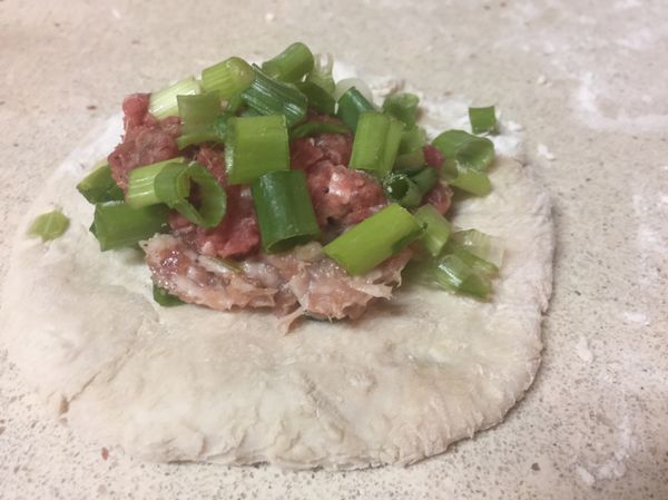 雙手沾麵粉，開始放餡料：球團沾滿粉後搓揉壓扁，放上絞肉及蔥花，包成球狀後，餡料包夾處朝下放，壓扁放盤子內，分兩盤。豬絞肉那盤我會放上芝麻做區分（餅上要擦一層油再放芝麻）。