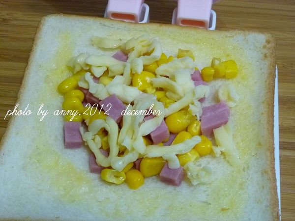 取一片土司片，塗上一層蛋液後，在土司中央放入1大匙的火腿丁.1大匙玉米粒及1/2大匙的起司絲，蓋上另一片塗上蛋液的土司片~