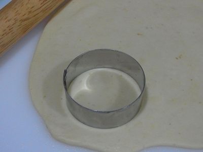 檯面上抹點油、擀薄麵糰，用圓型壓模器壓出一個圓形；另外作法：檯面灑點粉，麵糰分割小塊、滾圓、擀成圓形薄片。