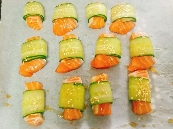 小黃瓜薄片沾上少許醬汁，將鮭魚捲起來，最後接口處朝下放在烘焙紙上，讓鮭魚卷不容易散開，最後表面撒上白芝麻。