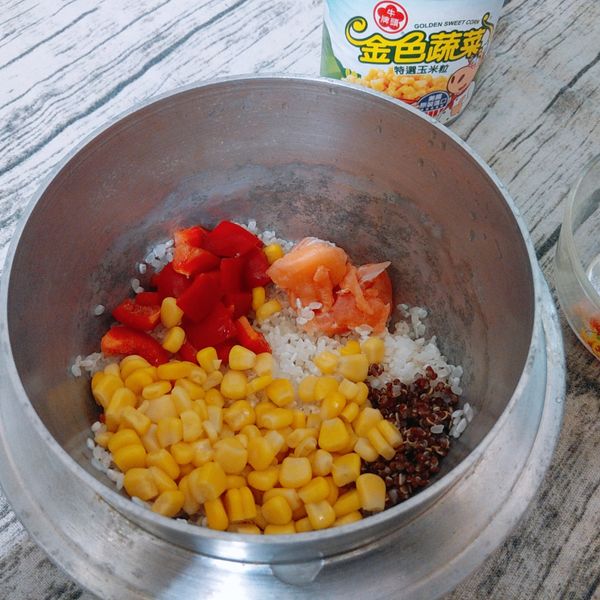 白米和紅藜麥分別清洗乾淨並瀝乾水份後放入鍋中，依續放入牛頭牌金色蔬菜特選玉米粒、鮭魚塊、紅甜椒丁，攪拌均勻備用。