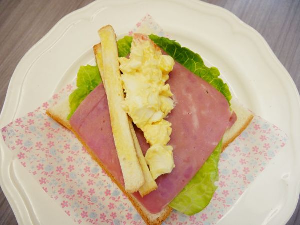 接著擺上火腿、烤過的去邊麵包條，把捏捏蛋沙拉捏一捏後擠入吐司上、接著左右往內捲並用油紙包起來就完成。