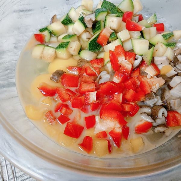 將雞蛋打入玻璃碗中，接著放入紅甜椒丁、黃甜椒丁、炒香的鮮香菇丁、炒軟的櫛瓜丁，加入雞粉和白胡椒粉一起攪拌均勻。