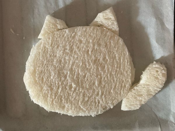 把吐司片剪出一個鵝蛋形做貓咪頭後，多出來的吐司剪出一個長的水滴型做貓咪手，再剪兩個小三角做耳朵