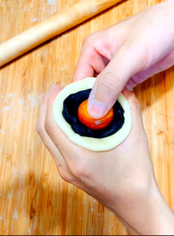 包入餡料:
將油皮收口處朝上，用手指從中間壓下去，將兩端接起來。
再將收口處朝下，用手掌稍微壓平，用擀麵棍上下左右來回桿一次，此時的油皮會變成圓形的形狀，底面為包餡的面，將剛剛備用的烏豆沙+蛋黃包入。