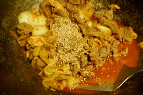 倒進韓式泡菜、鴻喜菇繼續翻炒，炒至稍稍收汁，稍微試一下味道，再酌量以鹽、糖稍稍[調味]，起鍋前加入白芝麻、蔥綠略為拌炒即完成