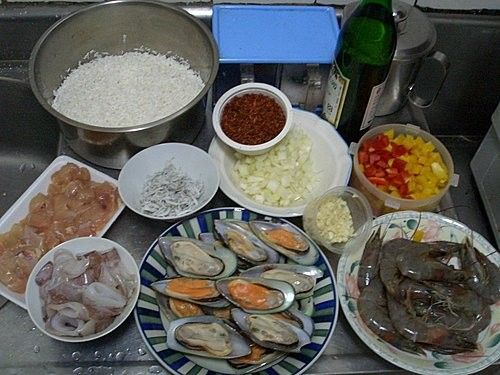首先準備材料，包括米、墨魚、蝦、淡菜、雞丁、紅黃椒、大蒜、洋蔥丁、我自己的做法還加了些小銀魚增加鮮味，另外要準備白酒提味用。鍋子記得要用平底鍋（材料見圖）