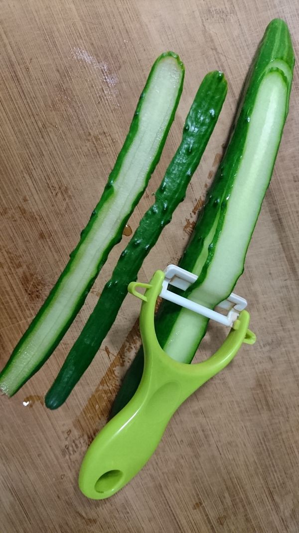 將小黃瓜放在帖板上，一手壓著小黃瓜，另一手用削皮刀將小黃瓜刨成片狀。