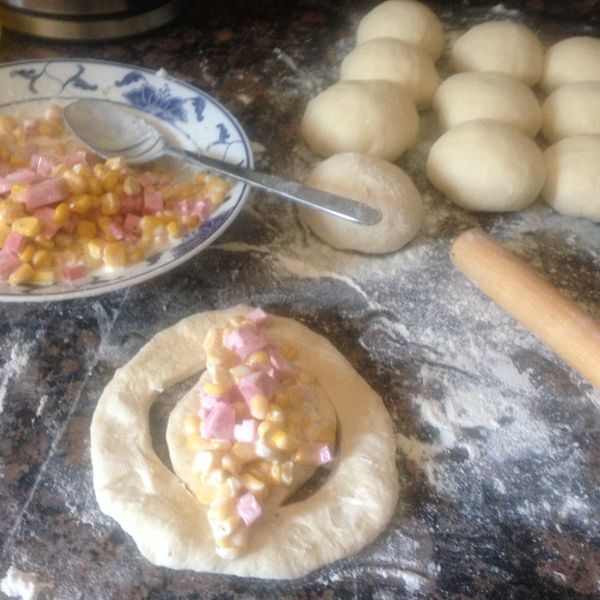 麵團發好以後，在工作台上倒些麵粉，將麵團倒出來揉麵整型，揉成長條以後，切成12個左右，每一個都整型再捏好成圓形的小麵團，12個都做好以後，不用再等，直接做。取其中一個麵團，撒些手粉，用桿麵棍桿成圓形，上面用披薩刀或是小刀刻兩個弧形刻痕如圖，中間放入玉米火腿內餡，如圖。